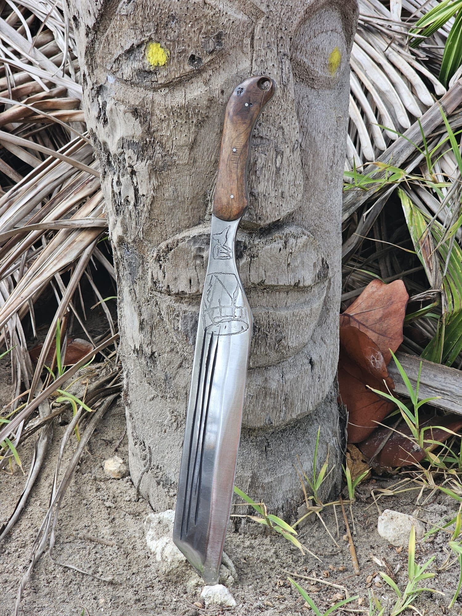 Sculpture de visage taillée dans un tronc de cocotier, une machette artisanale plantée devant, en bord de mer